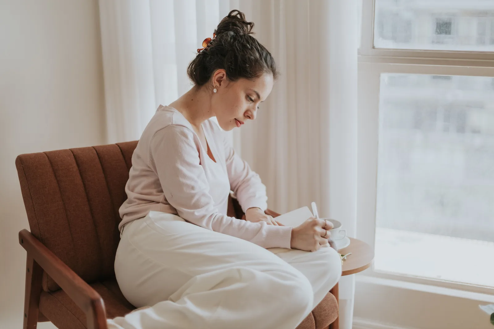 An adult woman writes in a notebook by a window.